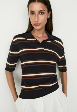 Donna che indossa una polo nera con strisce orizzontali marroni e beige e pantaloni bianchi, in piedi davanti a uno sfondo chiaro uniforme.