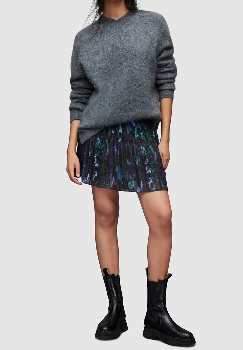 Femme portant un pull gris oversize, une mini-jupe plissée florale foncée et des bottes en cuir noires mi-mollet, debout devant un fond uni.
