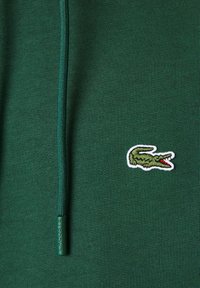 Tejido verde oscuro con un cordón verde y un logo de cocodrilo bordado en verde con la boca roja contorneada en blanco.