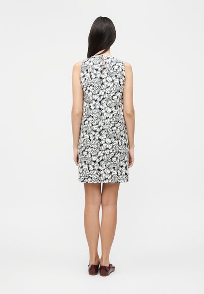 Γυναίκα με μακριά σκούρα μαλλιά, που φοράει αμάνικο ασπρόμαυρο λουλουδάτο φόρεμα και καφέ επίπεδα παπούτσια, στέκεται με την πλάτη σε λευκό φόντο.