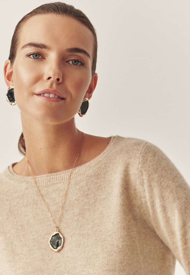 Kuldtooni ehtekomplekt, mis sisaldab suurt musta kalliskivi ripatsit ja sobivaid kõrvarõngaid. Modell kannab beeži kampsunit ümmarguse kaelusega.