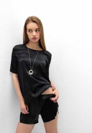 Junge Frau in schwarzem Satin-Top und Shorts mit beigen Spitzendetails, trägt eine runde Anhänger-Halskette, schaut seitlich an eine weiße Wand.