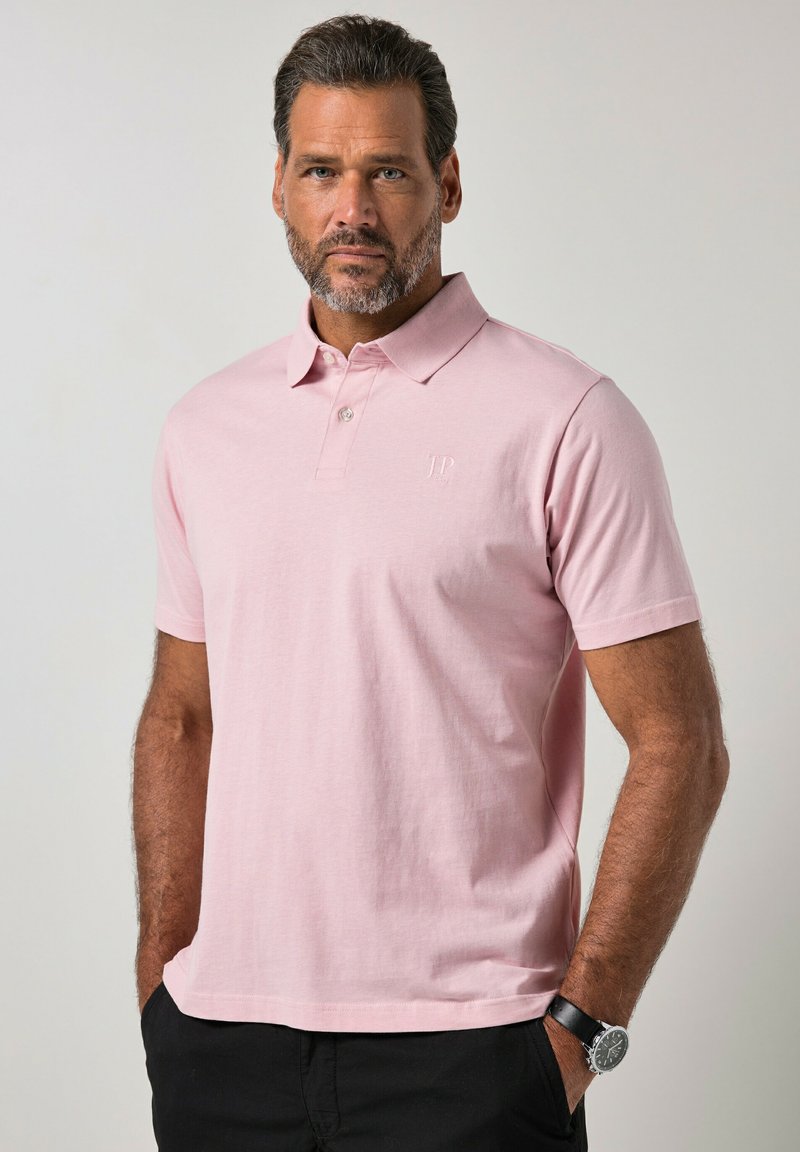 Hombre con barba que lleva una camiseta tipo polo de color rosa claro y pantalones negros, de pie con las manos en los bolsillos frente a un fondo liso.