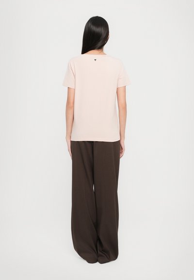 Light pink short-sleeve top with a round neckline and small logo detail at the back, paired with wide brown trousers for a relaxed fit.