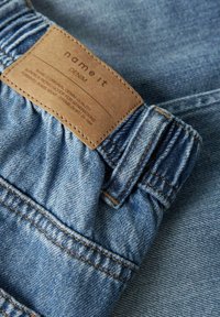 Tissu en denim bleu clair présentant une taille élastique et une étiquette en cuir marron embossée avec "name it" et "DENIM". Un gros plan montre les détails de la couture.