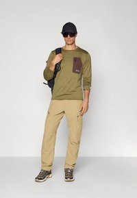 Άνδρας wearing ελιόχρωμο t-shirt με μακριά μανίκια, μπεζ παντελόνια cargo, παπούτσια πεζοπορίας, μαύρο καπέλο και γυαλιά ηλίου, κουβαλώντας μαύρο σακίδιο στον ώμο.