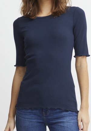 Vrouw draagt een aansluitend marineblauw geribd topje met geschulpte randen aan de mouwen en zoom, gecombineerd met medium gewassen blauwe spijkerbroek.