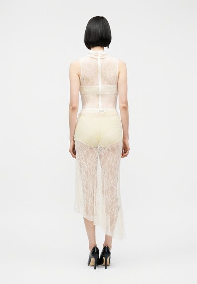 Femme aux cheveux courts noirs portant une robe blanche en dentelle transparente sans manches et des talons hauts noirs, debout de dos sur un fond blanc.