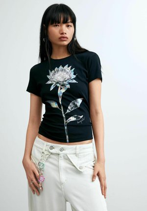 Ung kvinde iført en sort t-shirt med et stort holografisk blomsterdesign og hvide bukser, der står mod en ensfarvet lys baggrund.