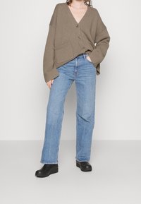 Khaki gestrickte Strickjacke mit V-Ausschnitt und drei Knöpfen, kombiniert mit hellblauen geraden Jeans und schwarzen Clogs. Taschen Details an der Strickjacke.