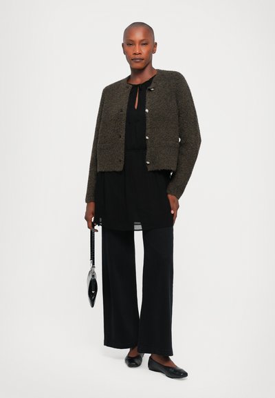 Knit jacket in dark green with silver button closure, paired with a black tunic dress and wide-leg black pants, holding a black handbag.
