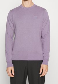 Jasnofioletowy sweter z długim rękawem wykonany z miękkiej tkaniny, z okrągłym dekoltem i małą naszywką z logo na piersi. Ściągacze na rękawach i u dołu.
