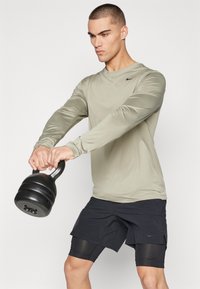 Langärmliges, hellolives Nike-Oberteil, schwarze Shorts, hält eine schwarze Kettlebell. Glatter Stoff, athletische Passform und minimale Markenkennzeichnung.