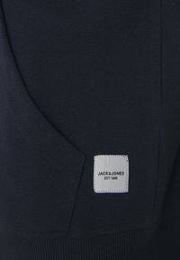 Námořnicky modrý svetr s klokaní kapsou, strukturální tkaninou a bílou obdélníkovou cedulkou s nápisem "JACK&JONES EST.1990" přišitým na něm.