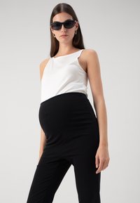 Tehotenský outfit s bielou, bezrukávovou blúzkou a čiernymi elastickými nohavicami navrhnutý pre pohodlie, s hladkou textúrou a priliehavým strihom.