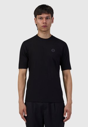 Young man standing facing forward wearing a plain black short-sleeve t-shirt with a small logo on the chest against a gray background.