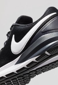 Scarpa sportiva nera con tomaia in rete, logo swoosh bianco, suola imbottita e battistrada in gomma nera testurizzata con dettagli di trazione.