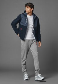 Heren hoodiejack, marineblauwe fleece met een zachte textuur, grijze stofpanelen, twee voorkezen, rode zippertrekken, gecombineerd met grijze joggingsbroeken en sneakers.