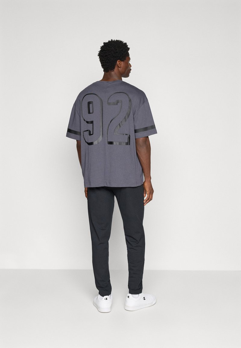 Graues übergroßes T-Shirt mit der schwarzen Zahl 92 auf dem Rücken, kombiniert mit dunklen Hosen und weißen Turnschuhen. Kurze Ärmel, lässige Passform.