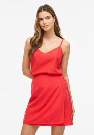 Femeie cu păr lung și șaten, purtând o rochie roșie fără mâneci, în fața unui fundal alb simplu.