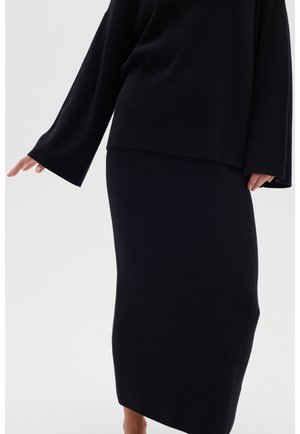 Person wearing a black long-sleeve top with wide sleeves and a matching black midi skirt against a plain light background.