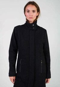 Manteau en laine noir avec un col montant, des poches latérales zippées et des détails de couture verticale à l'avant. Texture lisse et coupe ajustée.