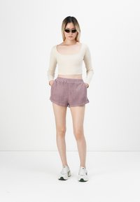 Haut beige à manches longues à côtes et shorts texturés mauves avec poches, associés à des baskets blanches avec des accents verts. Lunettes de soleil noires.