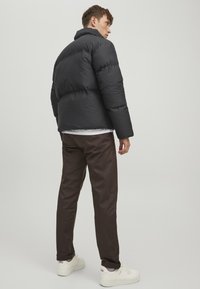 Schwarze Steppjacke mit hohem Kragen, ausgestattet mit gesteppten Abschnitten und einer glatten Textur. Kombiniert mit dunkelbraunen Hosen und weißen Turnschuhen.