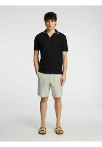 Črna polo majica z belim ovratnikom, v kombinaciji s svetlo zelenimi in belimi prugastimi kratkimi hlačami. Model nosi bež sandale in stoji z rokami v žepih.