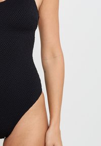 Gros plan sur le torse et le bras d'une personne portant un maillot de bain une pièce noir à texture, avec un petit tatouage sur l'intérieur de l'avant-bras.