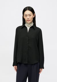 Crna košulja dugih rukava s prednjim kopčanjem na dugmad i ovratnikom, s glatkom teksturom i ravnim rubom, nosi se preko prugaste rolke.