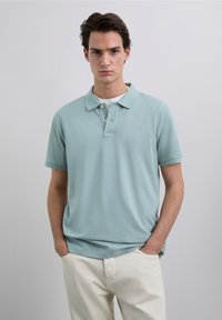 Tricou polo albastru deschis cu material texturat, mâneci scurte, guler clasic și trei nasturi, purtat cu pantaloni de culoare crem.