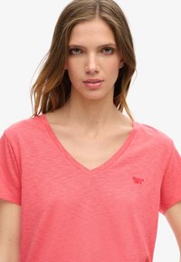 V-ringad kortärmad T-shirt i korallfärg med en mjuk textur. Har en liten röd logotyp på vänster bröst. Enkel design, inga mönster.