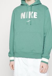 Zelená bavlněná mikina s kapucí a přední kapsou, se velkým bílým logem Nike. Obsahuje stahovací šňůrky a žebrované manžety s světelnými akcenty.