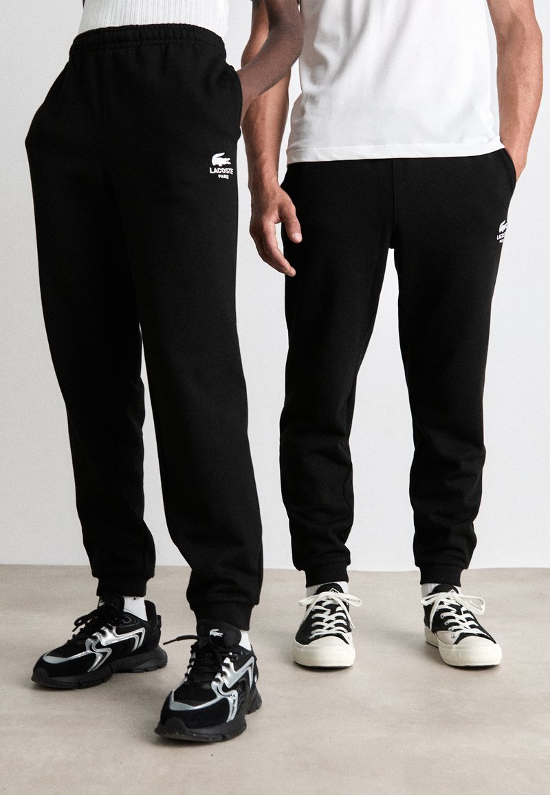 Zwarte sweatpants met elastische tailleband en boorden, voorzien van een geborduurd wit Lacoste-logo. Draag ze met contrasterende zwart-witte sneakers.