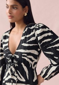 Robe noire et blanche à rayures zébrées avec un décolleté en V profond, une taille froncée et des manches bouffantes. Présente un détail à nouer au niveau du col.
