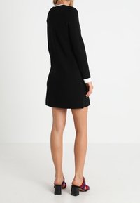 Žena v černých dlouhých rukávech mini šatech s bílými manžetami a vícebarevnými sandály na podpatku, stojící proti jednoduchému bílému pozadí.