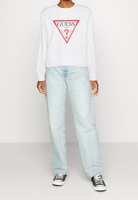 Persona che indossa un felpa bianca Guess con logo triangolare rosso, jeans azzurri chiari e sneakers nere e bianche, in piedi davanti a uno sfondo semplice.