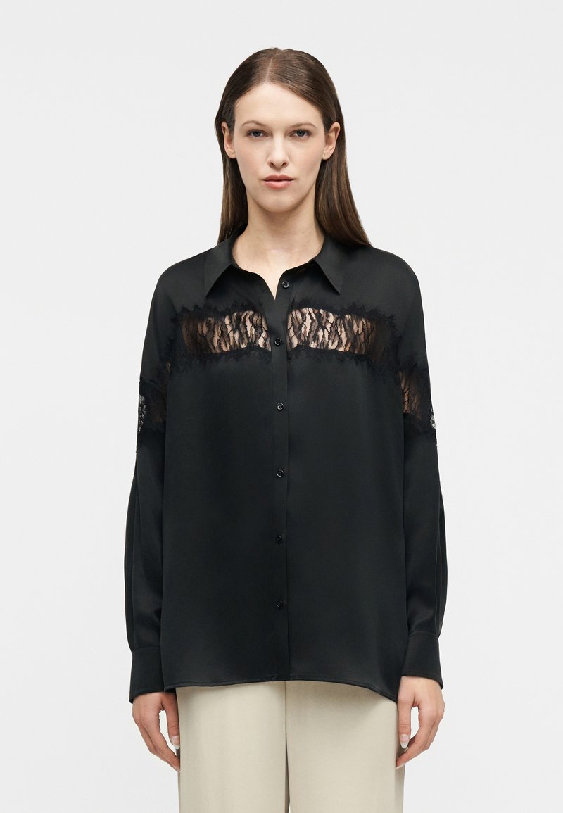 Γυναίκα με μαύρο πουκάμισο με μακριά μανίκια και κουμπιά, με δαντελένιο τμήμα στο στήθος και στα πάνω μανίκια, συνδυασμένο με ανοιχτό μπεζ παντελόνι.