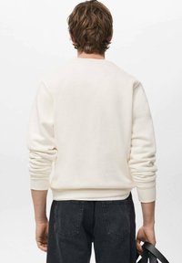 Sudadera color crema con puños y cintura acanalados, con un diseño sencillo y un ajuste cómodo. Combinada con vaqueros gris oscuro.