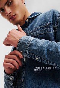 Tamsiai mėlynas džinsinis švarkas su mygtukų užsegimu, turintis apykaklę ir išsiuvinėtą logotipą "KARL LAGERFELD JEANS" priekyje.