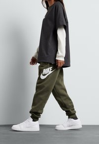 Tmavě šedé oversized tričko vrstvené přes bílé tričko s dlouhým rukávem, doplněné olivovými tepláky s velkým bílým logem Nike a bílými sneakers.
