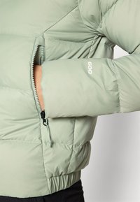 Lys grønn pufferjakke med glidelåslomme, elastisk midje og "600" logo detalj på ermet. Myk, quiltet tekstur.