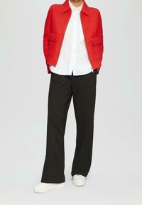 Červená bunda na zip s dvěma předními kapsami, bílá košile na knoflíky, černé kalhoty s širokými nohavicemi a bílé tenisky. Hladké textury a čisté linie.