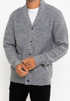 Homme portant un cardigan en maille texturée grise avec quatre boutons noirs sur une chemise blanche, mains jointes devant, pantalon noir visible.