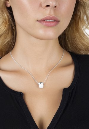 Gros plan d’une femme portant un collier en argent avec un pendentif rond blanc, vêtue d’un top noir à encolure boutonnée.