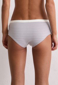 Kobieta od talii w dół, ubrana w jasnoszare majtki w cienkie białe paski poziome, stojąca na tle gładkiej białej ściany.