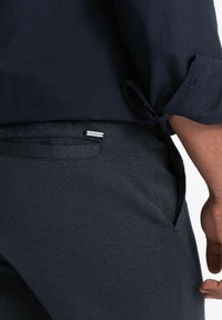 Camicia blu navy con maniche arrotolate indossata sopra pantaloni scuri con texture e un leggero motivo, con un piccolo accento metallico vicino alla tasca.