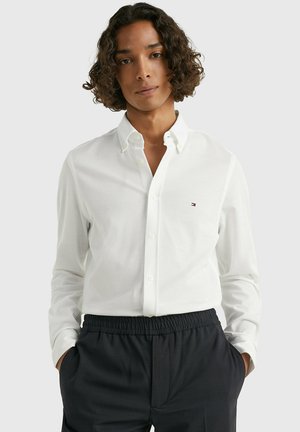 Camisa blanca de manga larga con botones y cuello, que presenta un pequeño logo en el pecho. Combinada con pantalones marinos de cintura elástica.