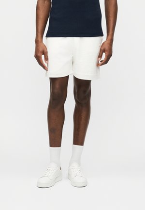 Moški v belih hlačah, belih nogavicah, belih supergah in črni majici stoji pred enobarvno svetlo ozadje.